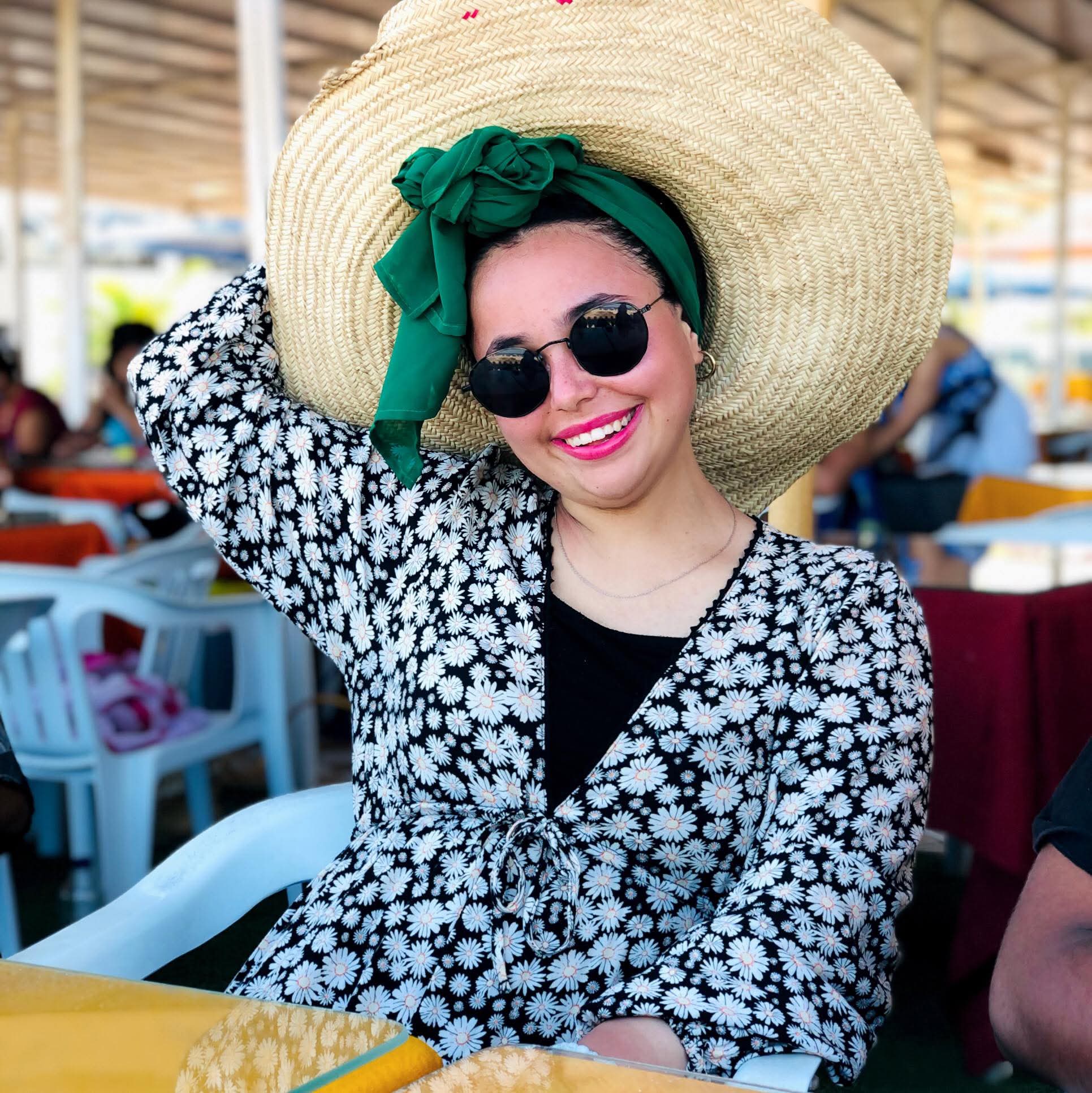 Lächelnde Frau mit riesigem Strohhut, grünem Kopftuch, Sonnenbrille und einem Kleid mit floralem Muster.