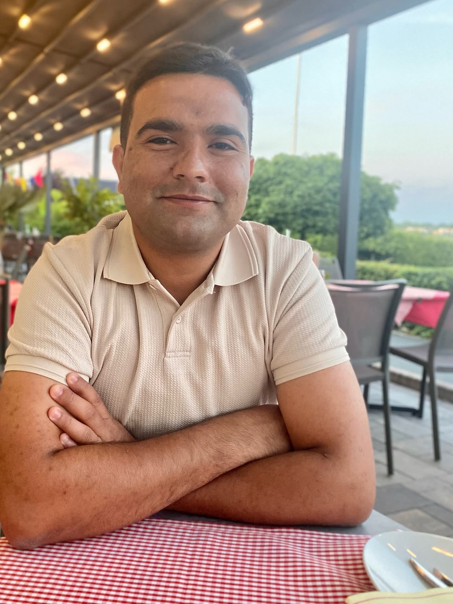 Lächelnder Mann im beigen Poloshirt sitzt mit verschränkten Armen an einem Tisch im Freien.