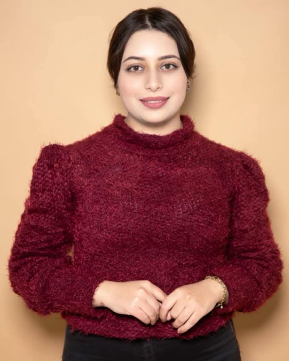 Lächelnde junge Frau mit dunklem Haar in einem flauschigen burgunderroten Pullover vor beigem Hintergrund.