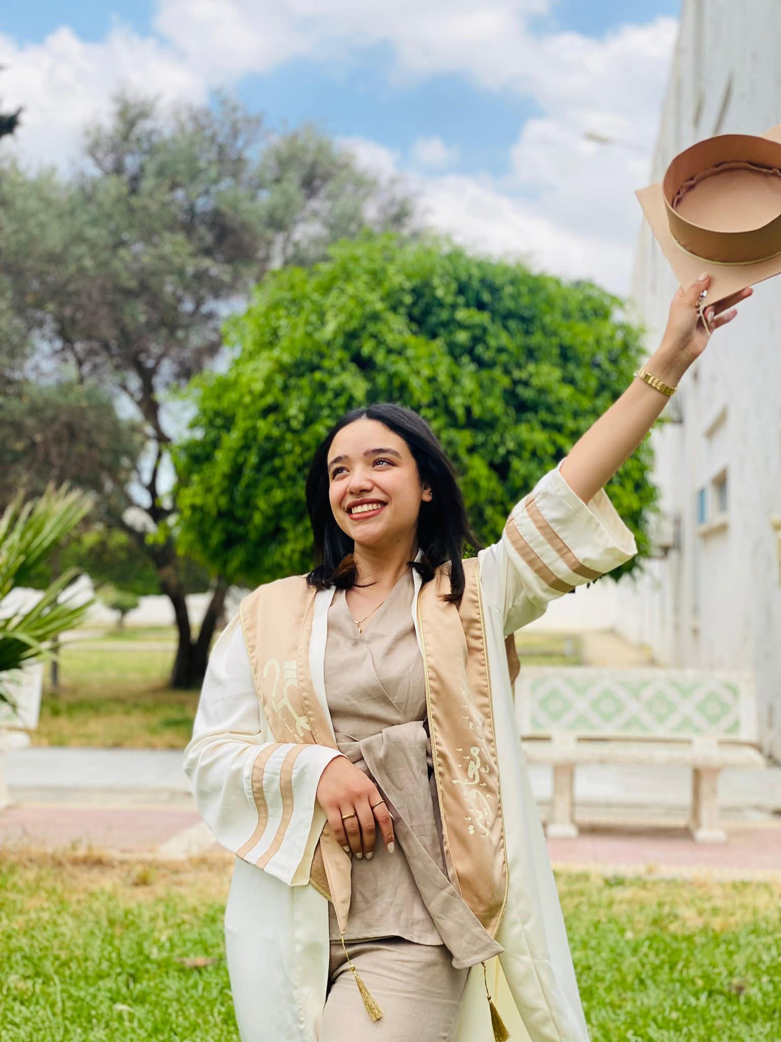Lächelnde Absolventin in weißer Robe hält ihren Doktorhut im Freien triumphierend in die Höhe.