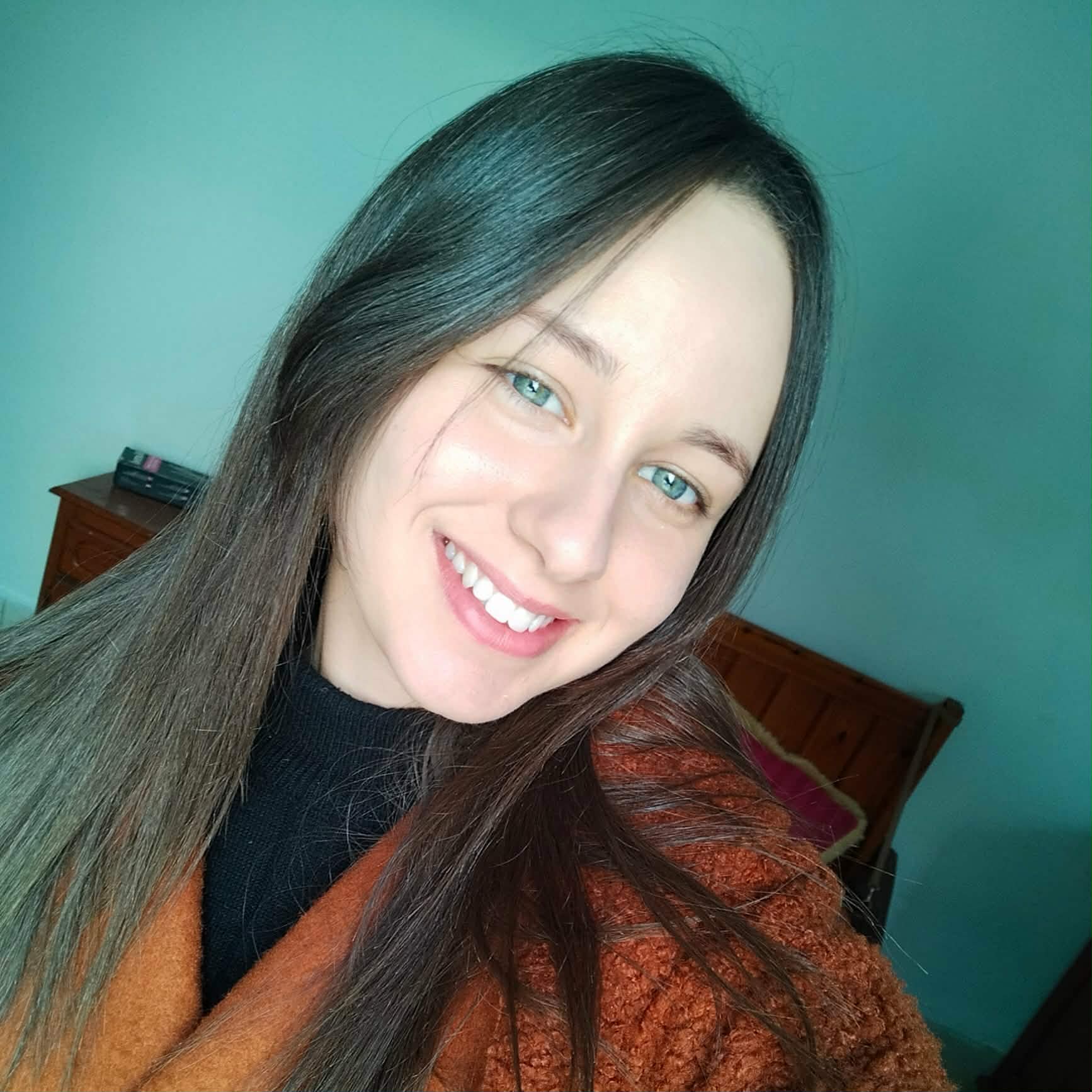 Lächelnde junge Frau mit langen dunklen Haaren und grünen Augen in einem braunen Mantel.