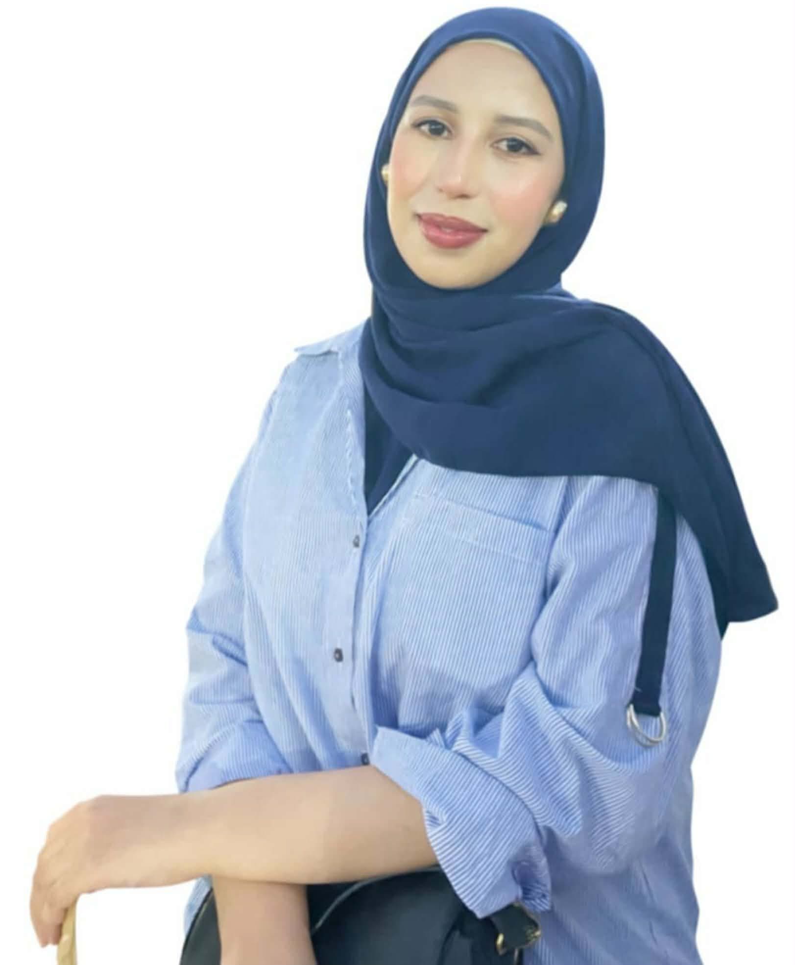 Lächelnde Frau mit dunkelblauem Hidschab und hellblau gestreiftem Hemd vor einem weißen Hintergrund.